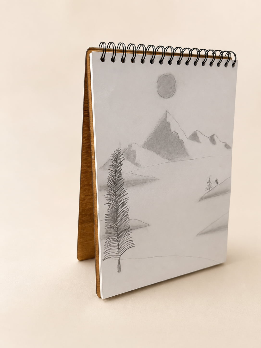 A spiral-bound notebook with a light gray cover featuring a minimalist pencil sketch of mountains, a tree, and a sun, displayed on a wooden stand against a warm beige background.