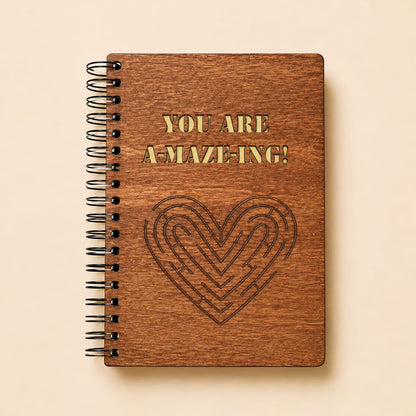 A spiral-bound notebook with a brown wood-grain cover, featuring the text 'YOU ARE A-MAZE-ING!' in gold and a dark engraved maze heart on a light beige background.