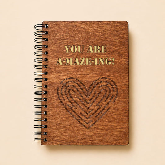 A spiral-bound notebook with a brown wood-grain cover, featuring the text 'YOU ARE A-MAZE-ING!' in gold and a dark engraved maze heart on a light beige background.