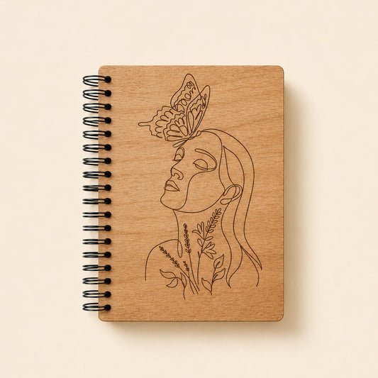 A spiral-bound notebook with a light natural wood cover featuring a black line art illustration of a woman's profile with a butterfly on her head and botanical details. The notebook is photographed flat against a solid, warm cream background.