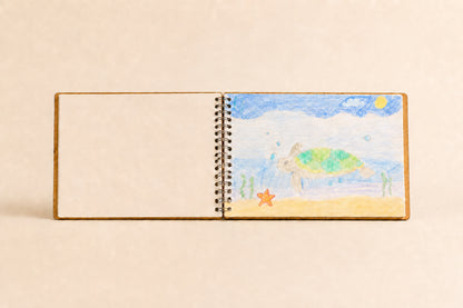 A small spiral-bound sketchbook or notebook standing upright on a beige background. The left page is blank, while the right page shows a colorful painted beach scene with a sea turtle, blue sky, yellow sun, and sand.