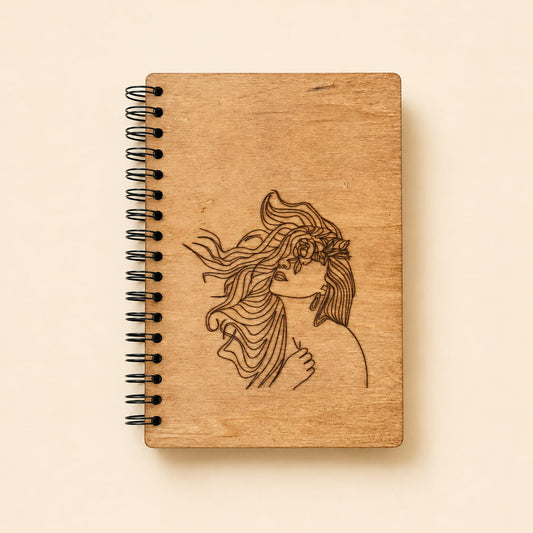 A small spiral-bound notebook with a natural wood cover featuring a dark engraved illustration of a woman with flowing hair, shown on a warm beige background.