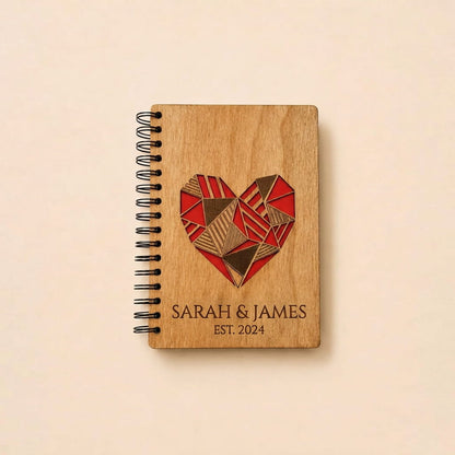 A personalized wooden spiral notebook with a black wire binding, featuring a red and brown geometric heart design and the engraved names 'SARAH & JAMES' and 'EST. 2024' on a light beige background.