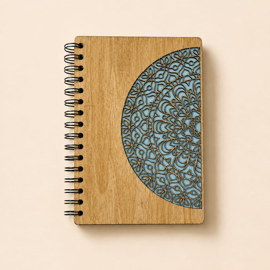 A spiral-bound notebook with a light wood cover featuring a laser-engraved blue mandala design on the right side, photographed on a solid beige background.