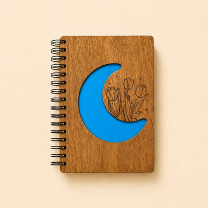 A spiral-bound wooden notebook with a laser-cut blue crescent moon and tulip design on the cover, photographed on a warm beige background.