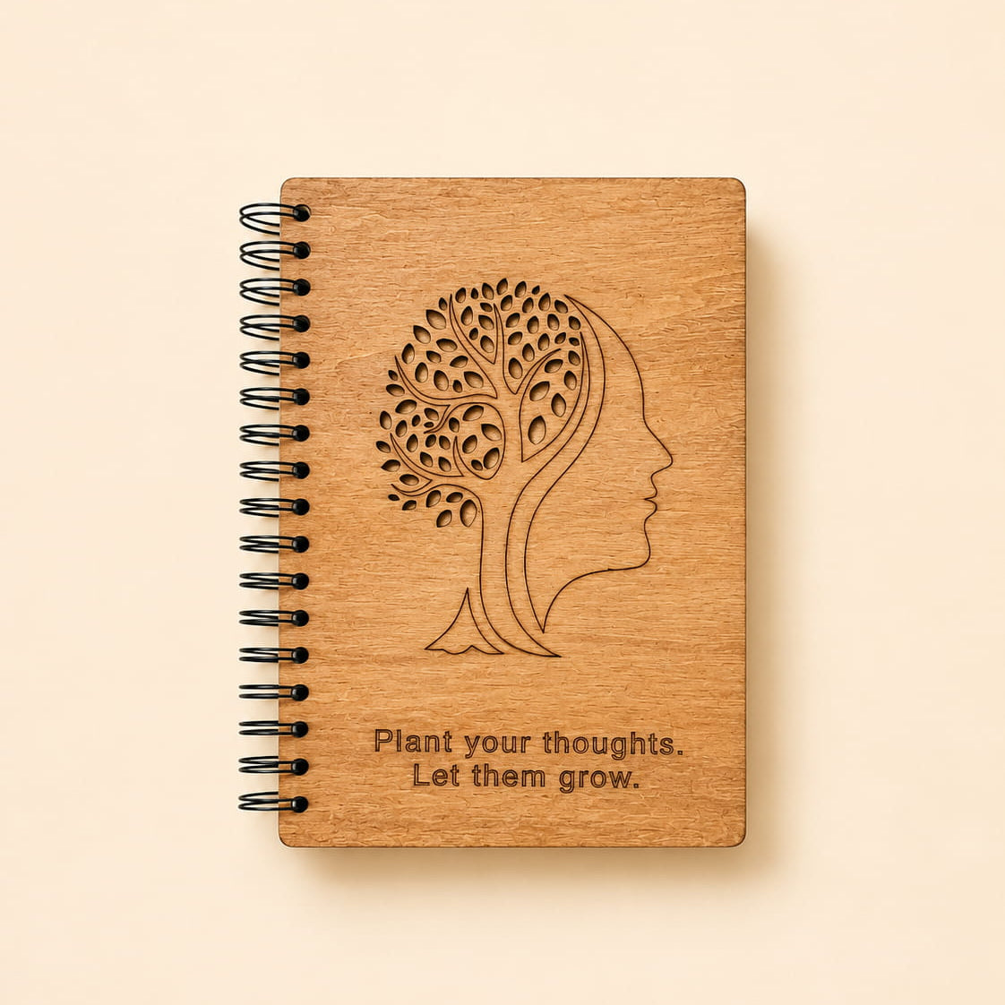 A wooden spiral notebook with a black wire binding, featuring an engraved tree-of-life head profile and the quote 'Plant your thoughts. Let them grow.