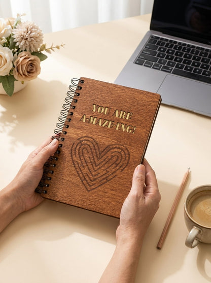 A person holds a spiral-bound notebook with a brown wood-grain cover that reads 'YOU ARE AMAZING!' and features an engraved heart design. The scene is set on a light beige desk with a laptop, flowers, a pencil, and a cup of coffee in the background.