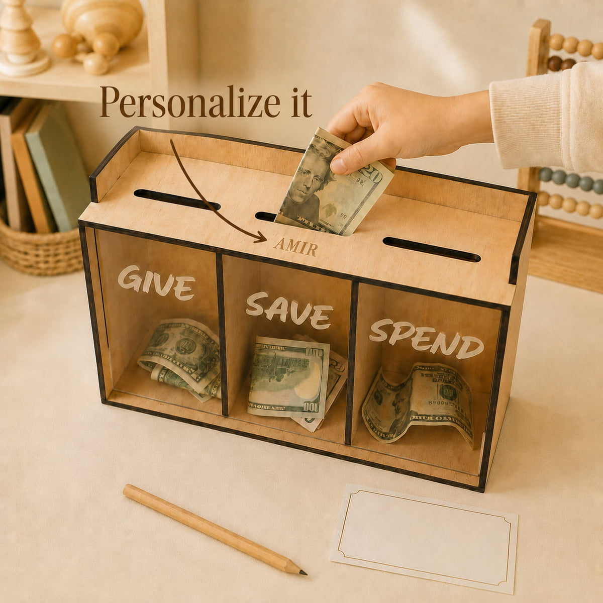 A wooden money box with three labeled compartments for GIVE, SAVE, and SPEND, shown on a tabletop with cash being deposited into the top slot. A hand is visible, and the scene includes decorative background objects and text reading 'Personalize it' and 'AMIR'.