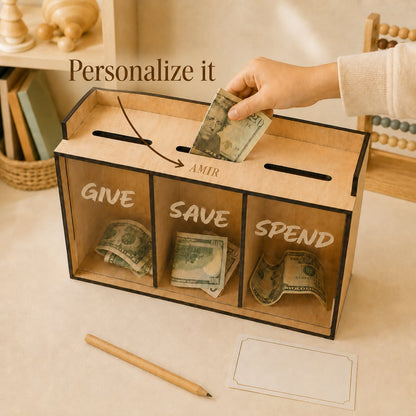 A wooden money box with three labeled compartments for GIVE, SAVE, and SPEND, shown on a tabletop with cash being deposited into the top slot. A hand is visible, and the scene includes decorative background objects and text reading 'Personalize it' and 'AMIR'.